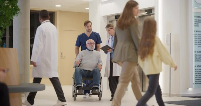Elderly Patient in Wheelchair Consulting with Female Doctor and Male Nurse in Busy Hospital Lobby as They Exit the Elevator. Medical Staff Showing Medical Information on Tablet Computer