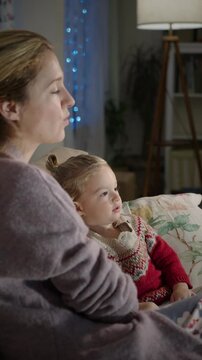 Young woman with her toddler sitting in a cozy living room watching TV in the evening. The room is softly illuminated by the glow of the television, highlighting the child's focused expression.