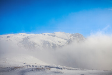 Obraz premium A breathtaking snowy mountain landscape shrouded in mist, with a vibrant blue sky above. The serene and mystical view captures the peaceful beauty of winter in high altitudes.