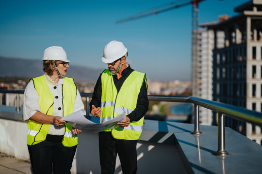 Two architects in safety gear review blueprints on a rooftop construction site, discussing project plans. The background showcases urban development. This image highlights teamwork, planning, and