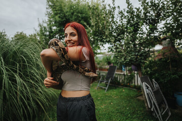 A girl with red hair smiles while holding a snake in a vibrant garden. The setting exudes a sense of adventure and connection with nature.