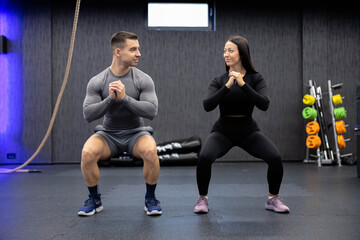A male and female athlete engage in a synchronized squat position within a gym, aligning perfectly...