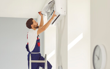 Professional electrician man with screwdriver maintaining, cleaning modern air conditioner indoors....