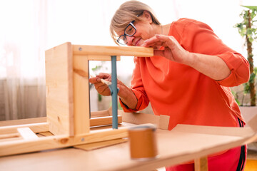 An elderly woman in orange casual clothes is painting a wooden chair in the living room