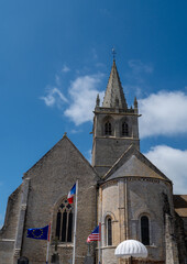 Obraz premium Large church with a flagpole in front of it. The flag is blue, white and red. The church is very old and has a steeple. Saint Come du Mont, Normandy, France
