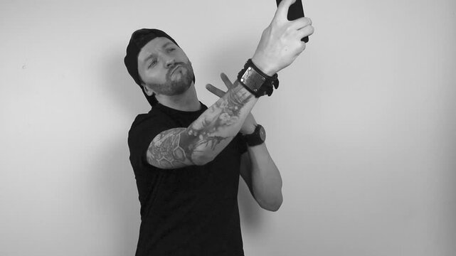 Grayscale footage of a tattooed man posing to a smartphone or phone camera and taking a selfie while showing a peace sign with his other hand. Man looks smug and happy to post on social media.