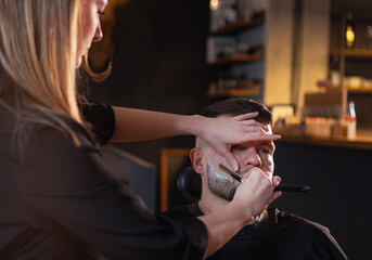 Long-haired young female Barber gently shaving bearded man face bristle while he in chair. Modern...