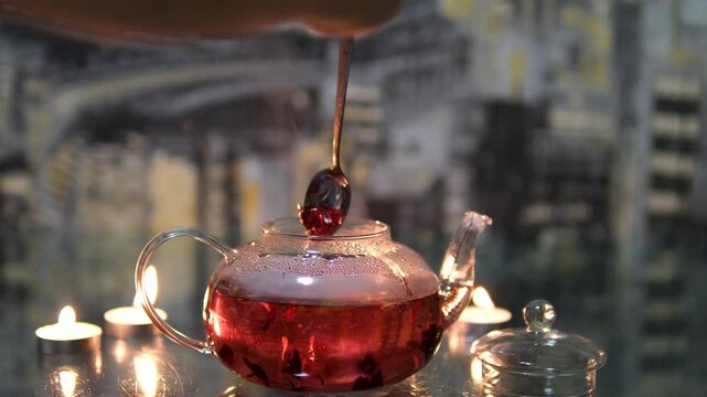 The process of making red hibiscus tea from sudanese rose flowers in a glass teapot. and stir red hibiscus tea with a spoon in a glass teapot on a dark background by candlelight