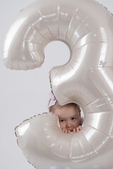 The image shows a young girl in a lavender dress embracing a sizable silver balloon in the shape of the number three.