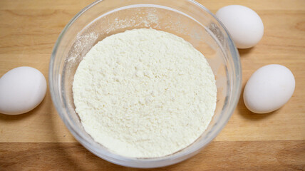 a bowl of flour stands on a wooden surface, three white chicken eggs lie nearby. cooking at home, homemade baking, following a recipe, preparing ingredients, kitchen tools.