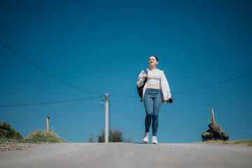 A girl confidently walks outdoors, wearing casual sportswear and headphones. She carries a backpack and enjoys the serene environment, embracing a sense of freedom and relaxation.