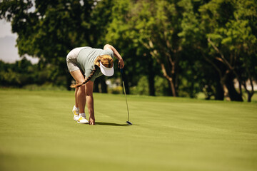 Woman golfer crouching on putting green with golf club preparing a shot in sunny weather - Powered by Adobe