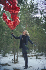 A young woman in a winter forest stands with heart-shaped balloons that soar upward. red balloons create a contrast with the green of the coniferous trees and the snowy . woman looks up with a dreamy