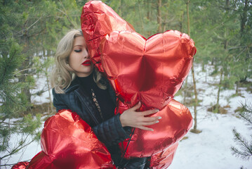 portrait of a young woman hugging red balloons in the shape of hearts . The girl with bright makeup and red lipstick holds the balloons to herself, creating an atmosphere of romance and tenderness.