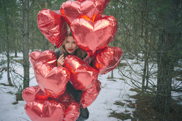 Romantic winter landscape with a woman holding a bunch of red balloons in the shape of hearts in a snowy forest among coniferous trees
