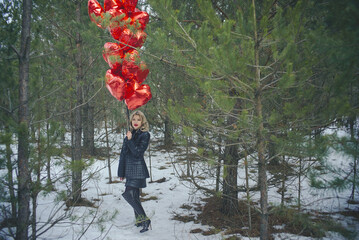A woman standing in a winter forest holding a bunch of red balloons in the shape of hearts. There is snow on the ground and the trees are surrounded by green coniferous branches.