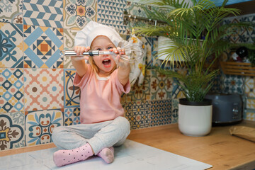A blonde Caucasian child girl toddler with a whisk in the kitchen sits on the countertop and helps...