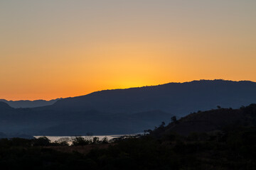Sunrise over Suchitoto, El Salvador, with a bright orange horizon, silhouetted hills, and Lake Suchitl&aacute;n in the foreground reflecting the morning light.