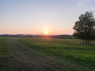 Orange sunset over green summer meadow with country road and forest with deciduous trees, clear sky background, Countryside landscape, copy space, Kokorin region, central Bohemia
