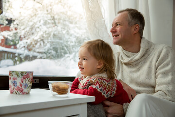 Middle age man and toddler sitting home in a chair by window with with scenic winter view of trees in snow. Bonding and quality family time together. 