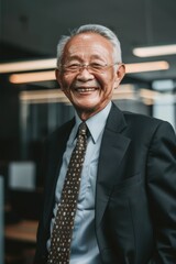 Naklejka premium Elderly Asian businessman wearing a dark suit and tie with a friendly smile. In an office environment, representing diversity and cultural competence in the corporate world.