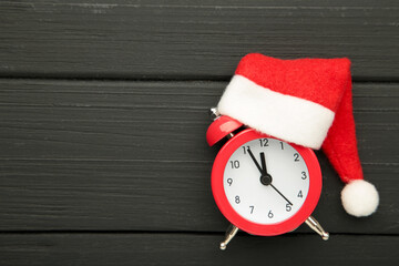 Red alarm clock with Christmas Santa hat on black wooden background. Top view.