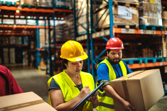 Warehouse workers in hard hats checking inventory and lifting boxes