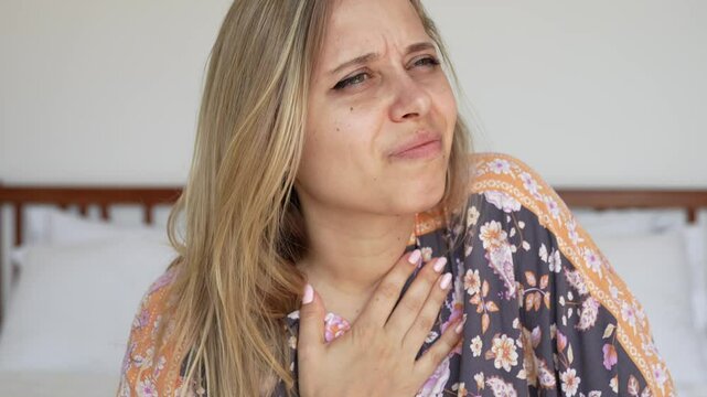Young caucasian blonde woman holding her chest with hand because of the nausea sitting on the bed in the room. Morning sickness, toxicosis, heartburn, poisoning, eating disorder