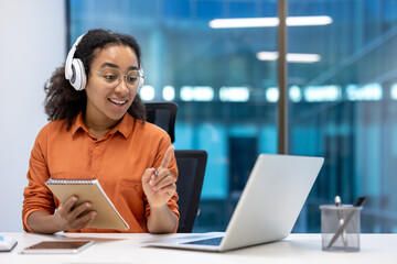 Young woman businesswoman participating in a virtual meeting while wearing headphones and smiling. She is seated at a desk with a laptop and notepad, focused and engaged in the discussion.