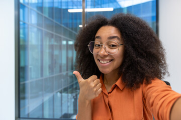 Young woman businesswoman in an orange shirt smiling and giving a thumbs up. She is in a bright,...