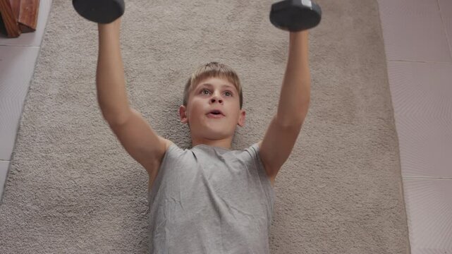 Young boy is training indoors, lying on a rug and lifting weights. He is wearing a grey t-shirt and is working out his arms