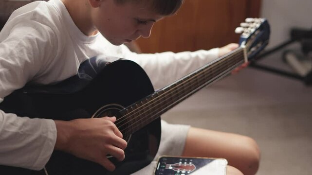 Young boy practicing acoustic guitar with a phone app. Carefully positioning fingers on the fretboard, he focuses on learning chords and developing musical skills at home