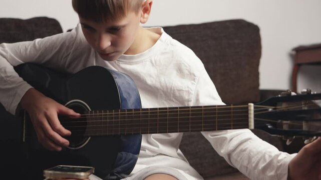 Young musician tuning a six-string acoustic guitar while seated on a sofa. Carefully adjusting the tuning pegs, he focuses intently on achieving the perfect sound