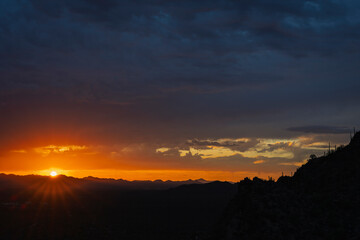 A breathtaking desert sunset casts vibrant colors over rugged terrain and dramatic skies, creating a serene and awe-inspiring scene ideal for nature enthusiasts.