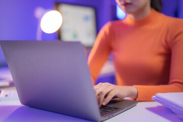 Young woman working from home at night typing on a laptop with a desk lamp and monitor in the...