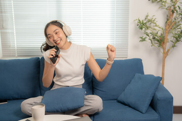 Obraz premium Sitting on a blue sofa in a living room, a young asian woman enjoys karaoke, using her smartphone as a microphone while singing and dancing joyfully