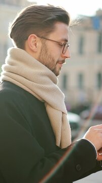 Internet addiction of modern people, young adult man using mobile gadget on city street. Surfing internet and chatting in social media, portrait of male person in coat and scarf standing in downtown