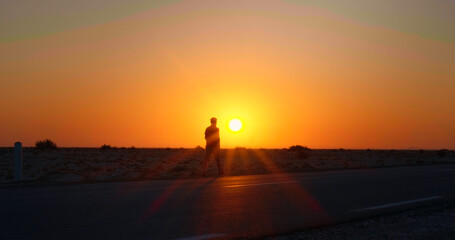 Fototapeta premium Man walking along the road with a sun set in the background. Sunset light in the dark and a man silhouette having a walk in sahara.