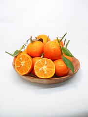 A wooden bowl filled with fresh mandarin oranges, some halved to showcase their juicy interior, accompanied by green leaves, set against a white background. Ideal for food or health concepts.  
