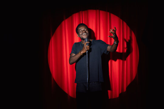 Smiling African American man speaking to microphone performing in comedy show open mike stand up on stage with red curtains.