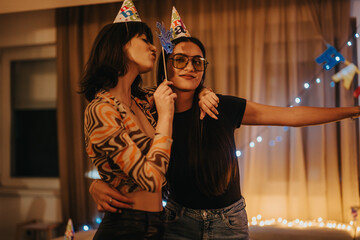 Two friends celebrating a birthday at home, embracing and wearing party hats. The warm, festive...