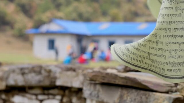 A yellow buddhist and tibet prayer flag is waving in the foreground. In the background there is a traditional house on a trekking path on the Himalayan mountain range
