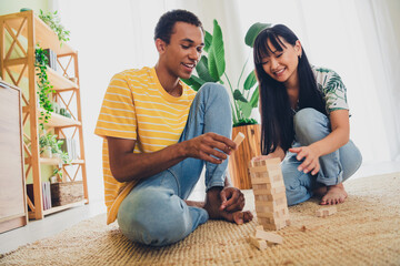 Photo of cute charming positive multinational couple sit floor play jenga build tower pile stack...