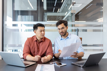 Two professionals engage in a discussion over important documents in office. Laptops and a phone are on the table, suggesting a tech-driven work environment. Collaboration and focus are evident.