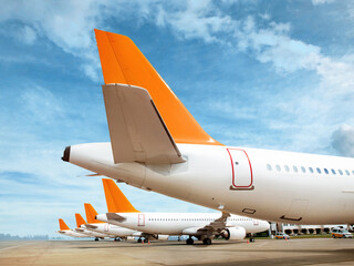 Multiple commercial airplanes lined up on the airport apron. Tail view with bright orange rudders under blue sky. Aviation and transportation concept.