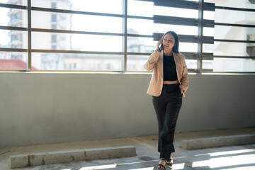 Fototapeta premium Young businesswoman is walking through a parking garage while talking on her phone, demonstrating the concept of connectivity and multitasking in a modern professional setting