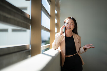 Fototapeta premium Smiling businesswoman talking on phone and gesturing with hand while leaning on wall near window in modern office building, enjoying conversation and bright sunlight
