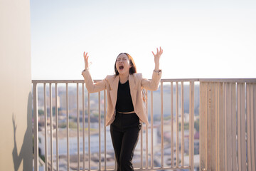 Businesswoman wearing a suit is standing on a balcony overlooking a city, screaming with her arms...