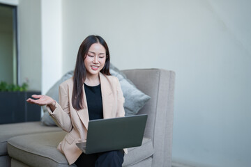 Young asian businesswoman in smart casual blazer using laptop for video conference, gesturing with hand while sitting on sofa in modern office lobby