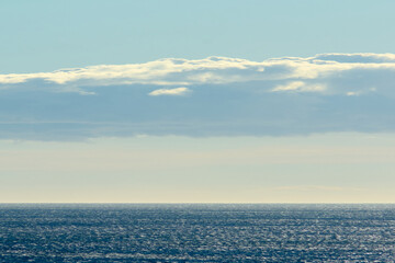 Fototapeta premium An expansive sea view with sunlight reflecting on the water. A large, illuminated cloud stretches across the sky, enhancing the serene and dynamic scenery.